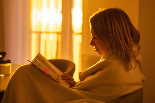 Smiling young beautiful woman reading a book, wrapped in a blanket while sitting in the living room at sunset light.