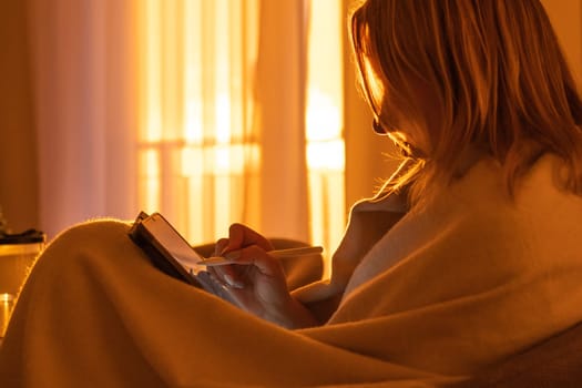A woman wrapped in a blanket and using a tablet while sitting in the living room at sunset light