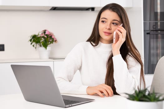 Female freelancer calls to friend on smartphone discussing life at break. Young woman talks on phone sitting at table near Notebook