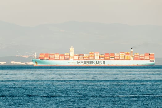 RIJEKA, CROATIA - JULY 10 2022: Container ship sails on deep blue ocean. White vessel transports containers against blurry dull mountain on coast and overcast sky on July 10 in Rijeka