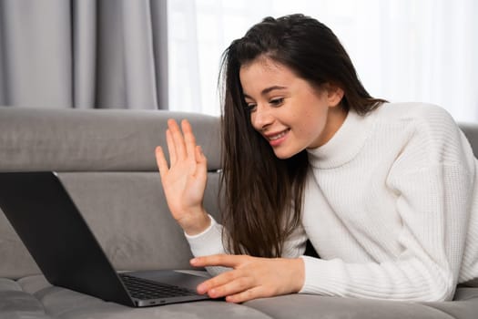 Brunette woman waves hello talking to boyfriend on video call via laptop. Young female person lies on comfortable sofa using modern gadget