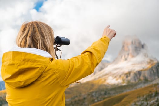 Blonde woman looks at Passo Giau pass through binoculars pointing finger on rocky mountain. Female tourist enjoys hiking in Italian Alps slow motion