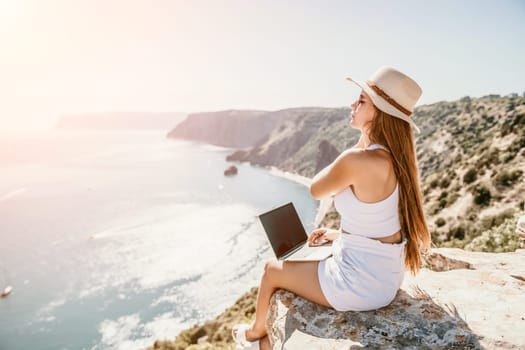 Successful business woman in yellow hat working on laptop by the sea. Pretty lady typing on computer at summer day outdoors. Freelance, travel and holidays concept.