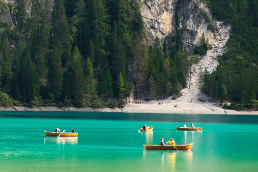 Tourists ride wooden boats on Braies lake with transparent water and Dolomites on the background in sunny day.