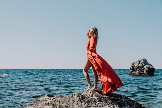 Woman travel sea. Young Happy woman in a long red dress posing on a beach near the sea on background of volcanic rocks, like in Iceland, sharing travel adventure journey