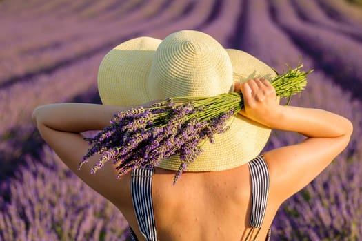 Woman in straw hat poses for photo holding bunch of fresh violet flowers. Young lady stands in lavender field on sunny day backside close view