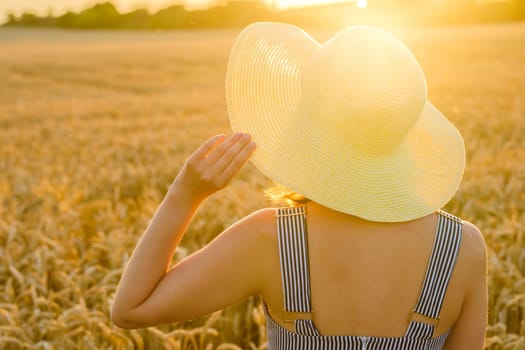 Woman holds on to straw hat standing in golden wheat field at bright sunset. Young lady poses for photo at rural site backside close view