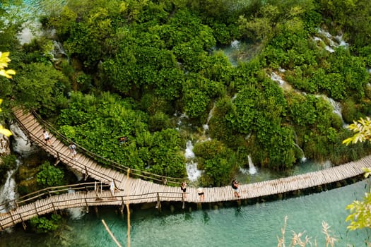 Hiking tourists walk on wooden footbridge surrounding lake in natural reserve. Majestic view of blue lake among picturesque landscape on Plitvice lakes upper view