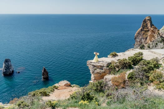 Woman in a yellow dress on the sea. Side view Young beautiful sensual woman in yellow long dress posing on a rock high above the sea at sunset. Girl in nature against the blue sky.