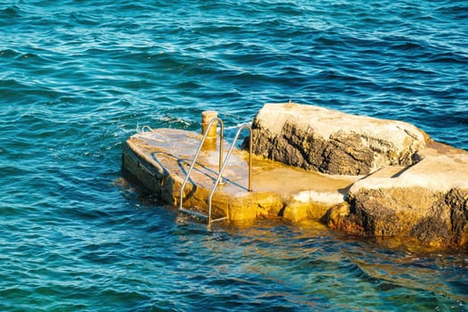 Metal ladder for swimmers descending into sea from small rock at sunlight. Stone pier washed by waves among deep turquoise sea at resort