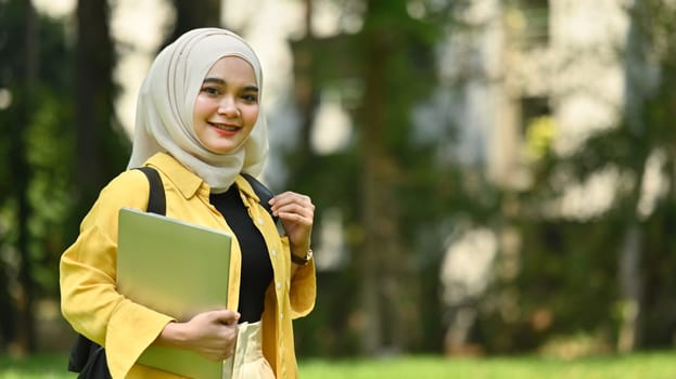 Smiling muslim college student holding laptop standing in the campus. Education, technology and lifestyle concept.