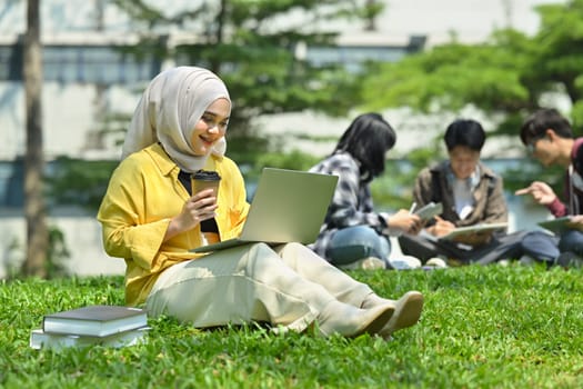 Smiling asian muslim student using laptop on green grass in campus with friends sitting on background.