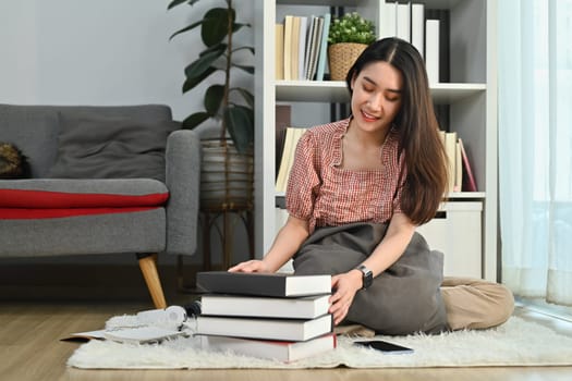 Full length of beautiful young woman sitting floor in bright living room with piles of books. People, leisure and lifestyle concept.