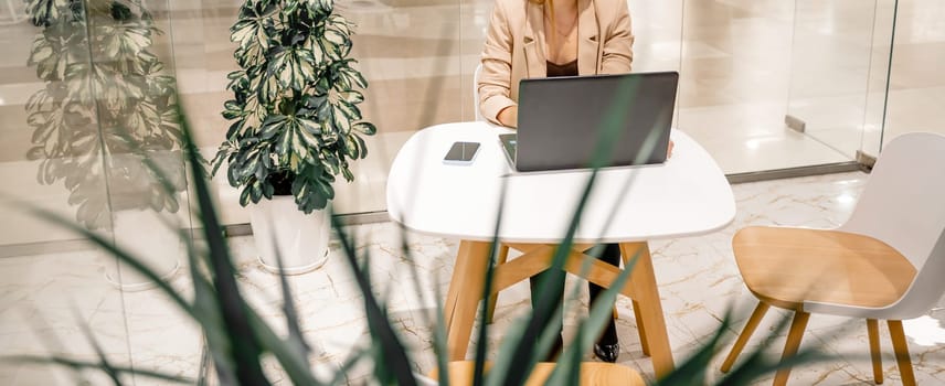 A business woman sits in a cafe and works at a computer. She is wearing a beige jacket