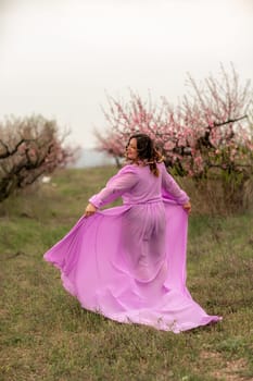 Woman peach blossom. Happy curly woman in pink dress walking in the garden of blossoming peach trees in spring.