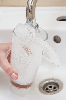 Female hand with glass. Glass is filled with transparent water under the tap with ceramic washbasin on the background