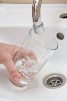 Female hand with glass. Glass is filled with transparent water under the tap with ceramic washbasin on the background