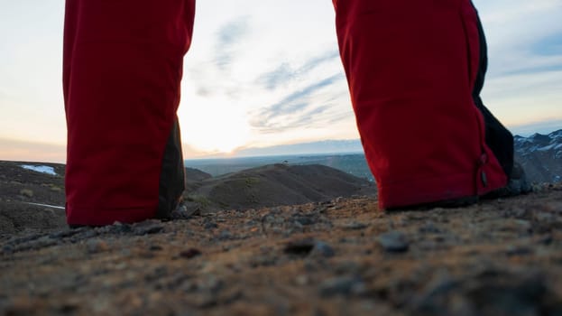 A guy in trekking boots takes steps through the Canyon. Stops at the edge. Sunrise. Orange-red rays of the sun and blue clouds. Red canyon land. Small stones. Sports red pants. View is like on Mars