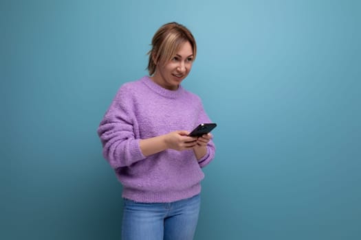 blond woman in purple hoodie chatting in smartphone on blue background.