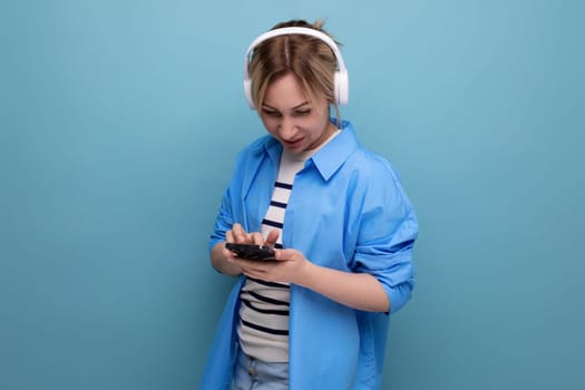 close-up photo of a bright smiling blond young woman dressed in a striped sweater and casual shirt with white wireless headphones communicates in the application in a smartphone on a blue background.