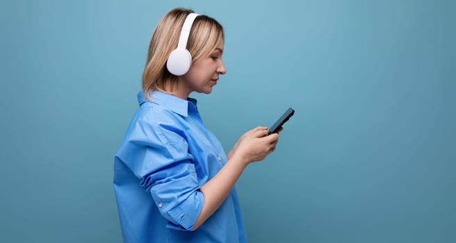 profile horizontal photo of an attentive millennial woman in a casual shirt with large white headphones with a smartphone in her hands on a blue isolated background with copy space.