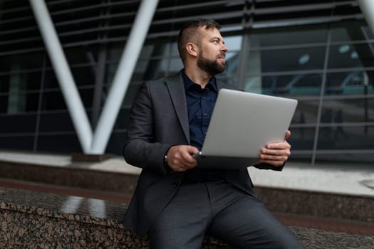 man in business suit sitting working on laptop outside.