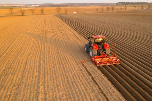 PRAGUE , CZECH REPUBLIC - MARCH 18 2022: Tractor drives at sunset leaving beds for planting vegetable seedlings in cultivated soil. Powerful machine operates on field at farm aerial view