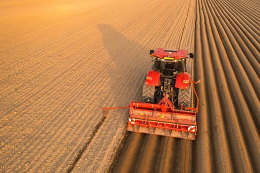 Tractor drags plow for leaving long furrows behind in soil on sunny day. Agricultural transportation works in endless field at sunset aerial view