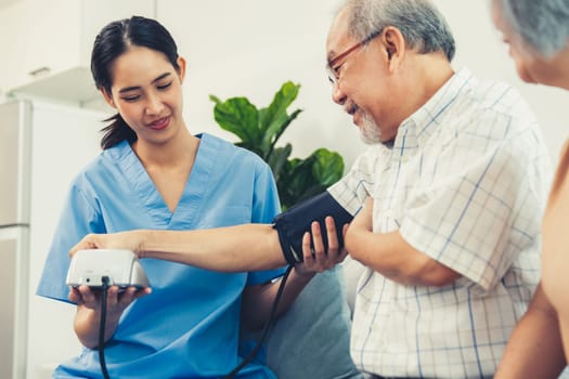 An elderly man having a blood pressure check by his personal caregiver with his wife sitting next to him in their home.