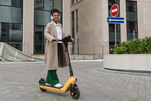 stylish african american woman rides an electric scooter against the backdrop of an office building.