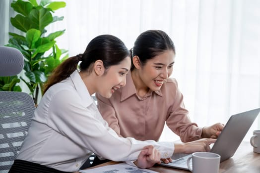 Two young office lady colleagues collaborating in modern office workspace, engaging in discussion and working together on laptop, showcasing their professionalism as modern office worker. Enthusiastic