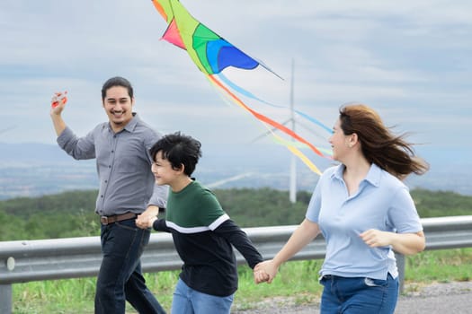 Progressive happy carefree family vacation concept. Young parents mother father and son run along and flying kite together on road with natural scenic on mountain and wind turbine background.