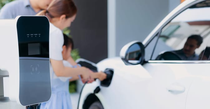 Closeup focus hand insert EV charger plug to electric vehicle at home charging station with blurred family in background. Progressive concept of green and clean energy to reduce CO2 emission by EV car