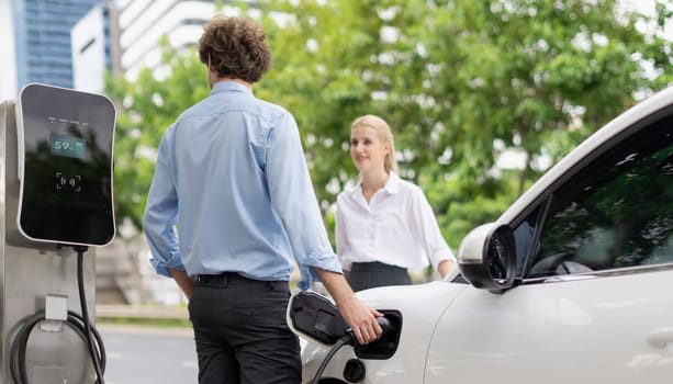 Progressive businessman and businesswoman with electric car parking and connected to public charging station before driving around city center. Eco friendly rechargeable car powered by clean energy.