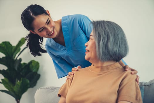 A caregiver rest her hands on the shoulders of a contented senior patient while she sitting on the sofa at home.