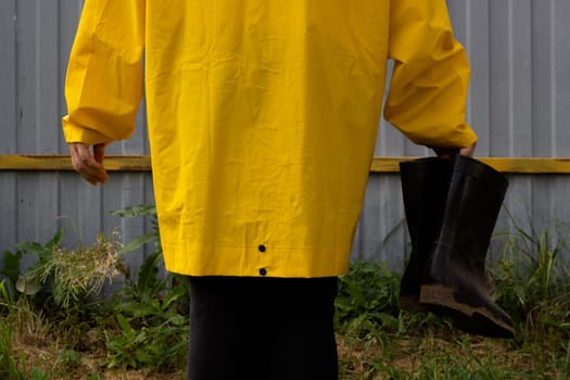 A woman in a yellow raincoat holds black rubber boots in her hand. Against the background of a gray metal fence.