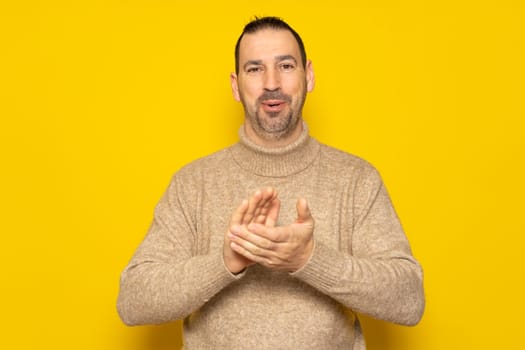 Bearded hispanic man wearing a beige turtleneck clapping heartily for good news, isolated over yellow background.