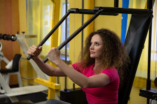 Workout performed at the gym. A woman uses sports equipment to exercise her upper body. Performing weight training exercises. Lifting weights at a gym. A well-equipped exercise room.