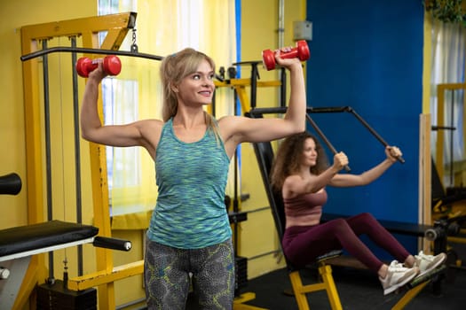 Two friends working out at the gym. In the foreground is a blonde woman lifting small red weights above her head. In the background, a girl with curly hair practicing on an exercise atlas