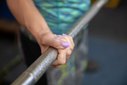 A woman's hand with purple painted nails. A woman clenches her hand while holding an exercise barbell in it. Lifting weights at a gym. Close-up of a woman's hand.