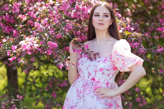 A beautiful brunette girl in a pink dress standing near pink blooming apple trees, holding a blooming branch, in the spring in the garden. Close up. Copy space