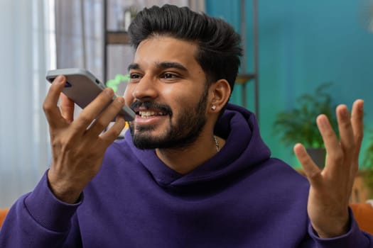 Happy excited indian man making online phone call conversation with friends. Portrait of hindu guy enjoying mobile loudspeaker talking chatting communication at home apartment living room on couch