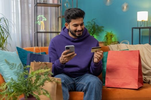 Happy shopaholic consumer indian man sitting with shopping bags at modern home apartment making online order, payment with credit plastic bank card. Discount sale on internet. Young hindu guy on sofa