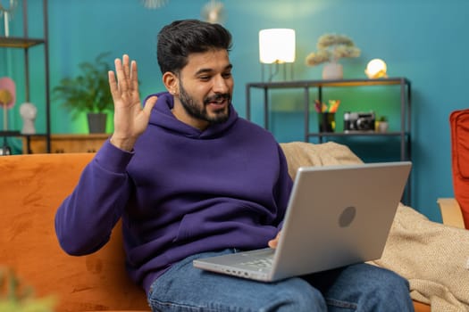 Indian man sitting on couch looking at camera, making video webcam conference call with friends or family, enjoying pleasant conversation. Young hindu guy laughing waving hello at home room