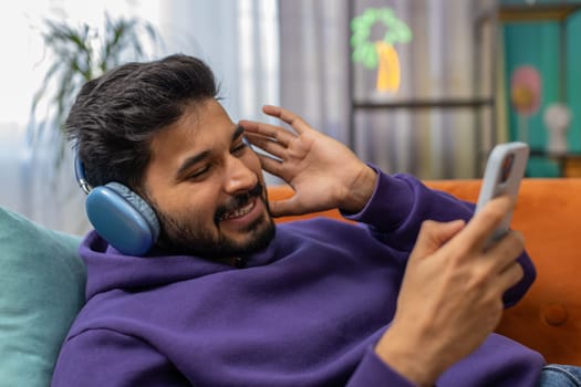 Happy overjoyed indian man in wireless headphones relaxing sitting on couch at home apartment choosing listening favorite energetic disco dancing music. People weekend daytime leisure activities