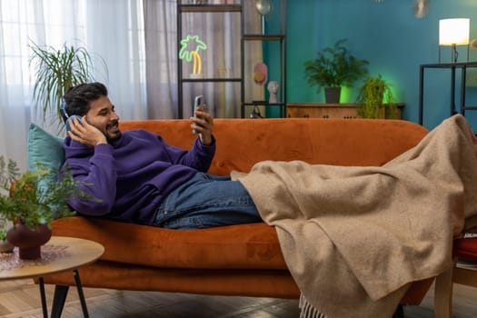 Happy overjoyed hindu indian man in wireless headphones relaxing sits on couch at home apartment choosing listening favorite energetic disco dancing music. People weekend daytime leisure activities