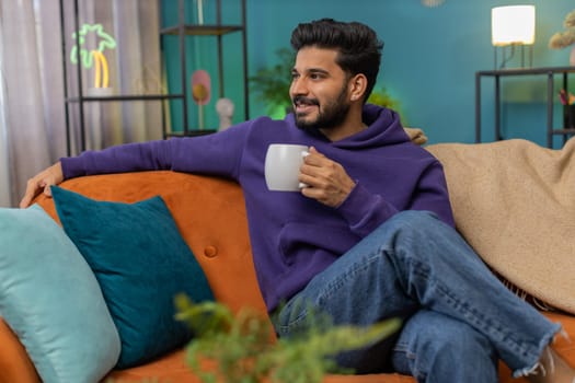 Handsome smiling indian man drinking a cup of warm coffee or herbal tea at home living room apartment. Break time. Hindu guy enjoying comfortable relaxing sitting on couch in the morning. Lifestyles