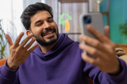 Happy indian man blogger taking selfie on smartphone, communicating video call online with social media subscribers followers, recording vlog stories. Young hindu guy at home apartment room on couch