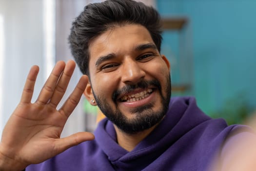 POV of indian man blogger taking selfie on smartphone, communicating video call online with social media subscribers followers, recording vlog stories. Young hindu guy at home apartment room on couch