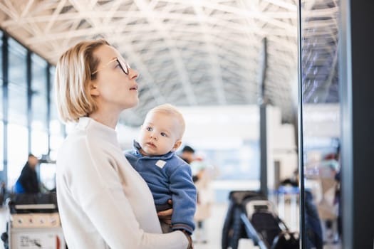 Mother traveling with child, holding his infant baby boy at airport terminal, checking flight schedule, waiting to board a plane. Travel with kids concept